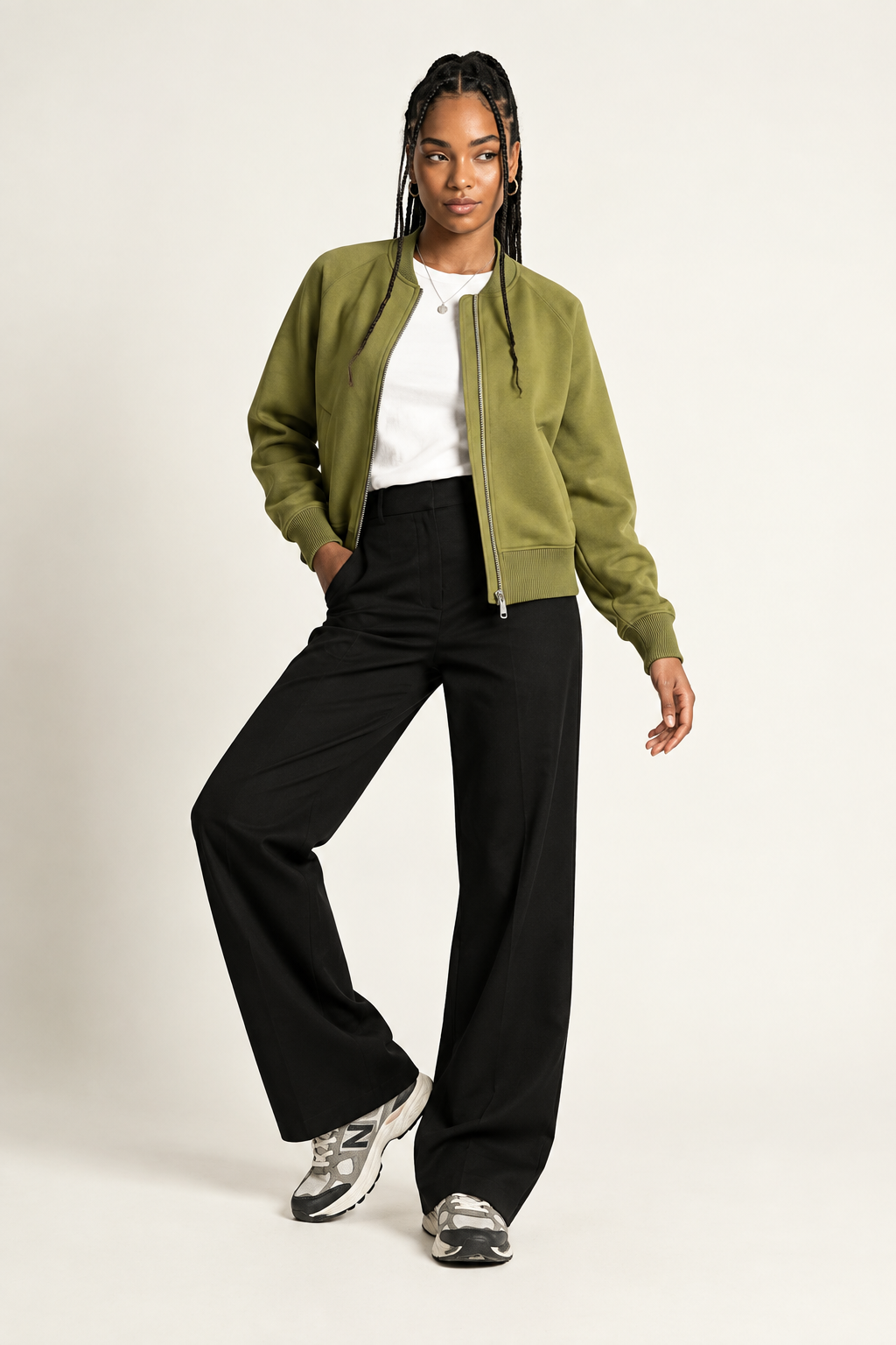 A model posing in a studio setting wearing an olive green bomber jacket, black wide-leg trousers, a white t-shirt, and gray sneakers against a light beige background.