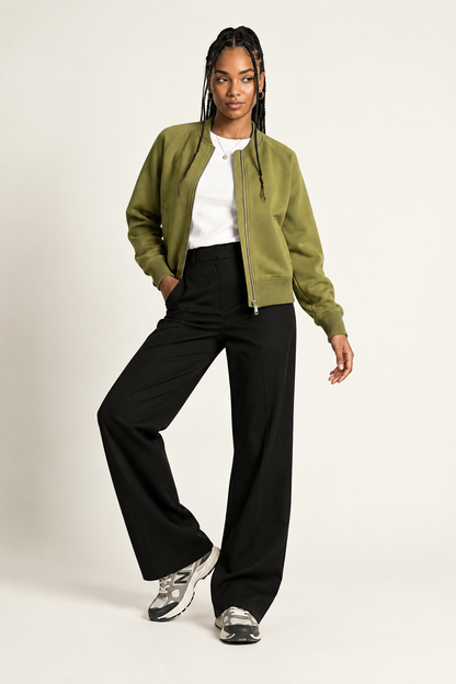 A model posing in a studio setting wearing an olive green bomber jacket, black wide-leg trousers, a white t-shirt, and gray sneakers against a light beige background.