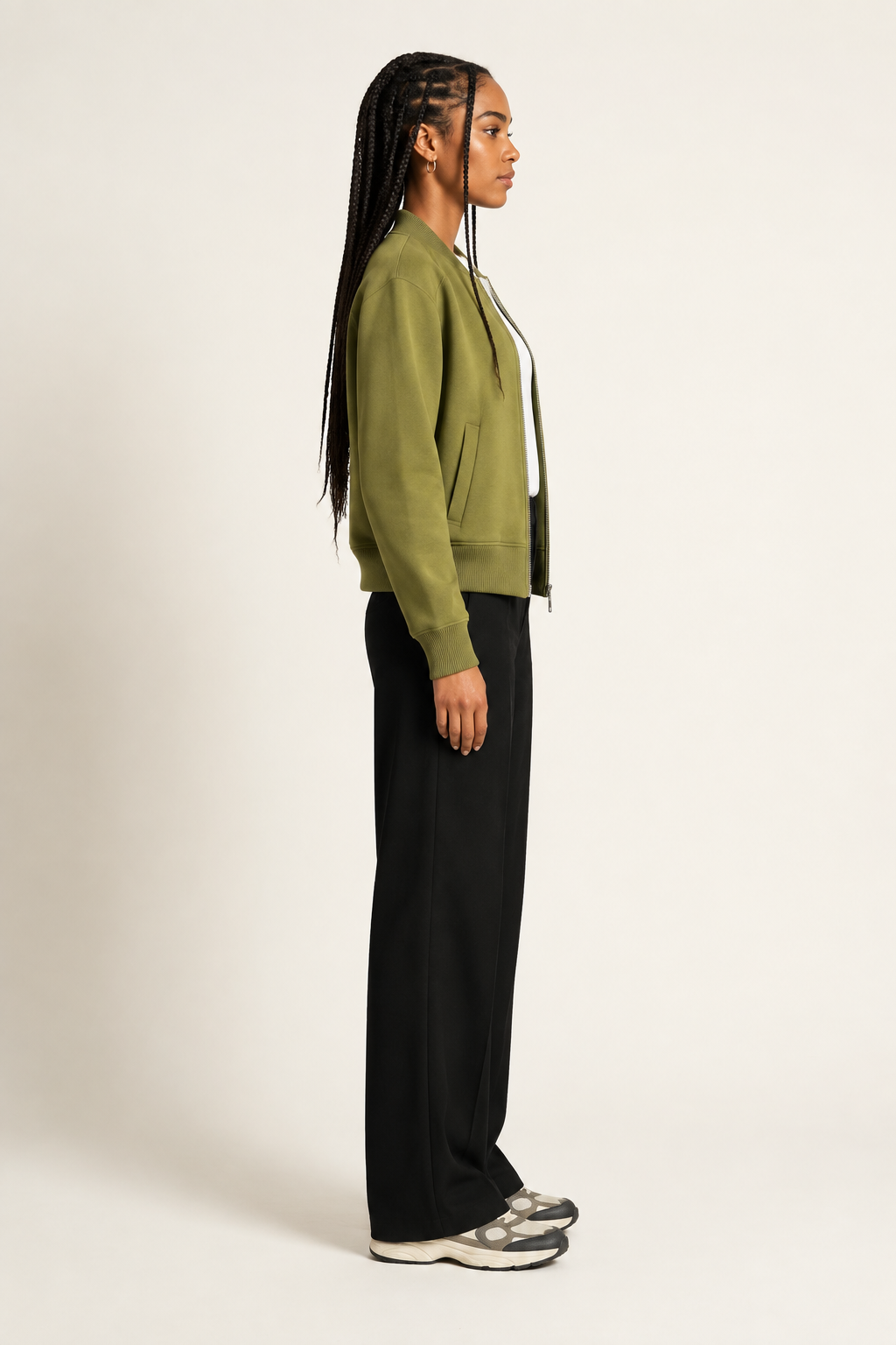 A woman in side profile wearing an olive green cropped zip-up jacket, black wide-leg trousers, and gray sneakers, standing against a plain off-white studio background.