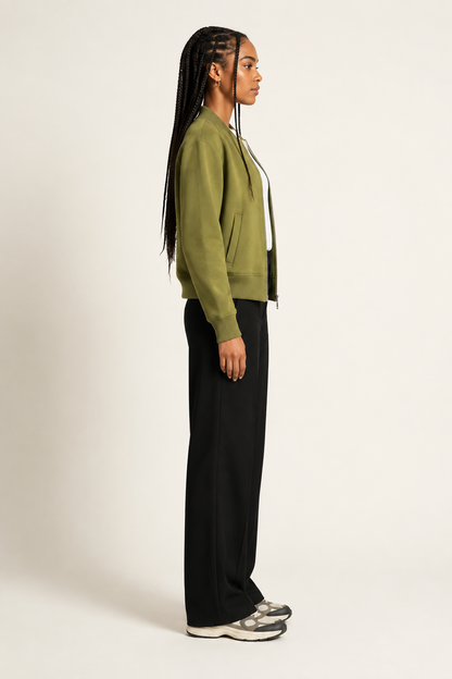 A woman in side profile wearing an olive green cropped zip-up jacket, black wide-leg trousers, and gray sneakers, standing against a plain off-white studio background.