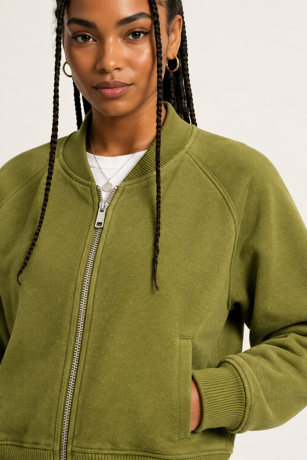 A close-up of a woman wearing an olive green zip-up bomber jacket over a white t-shirt, photographed against a clean off-white studio background.