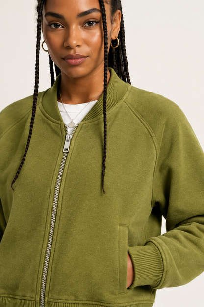 A close-up of a woman wearing an olive green zip-up bomber jacket over a white t-shirt, photographed against a clean off-white studio background.