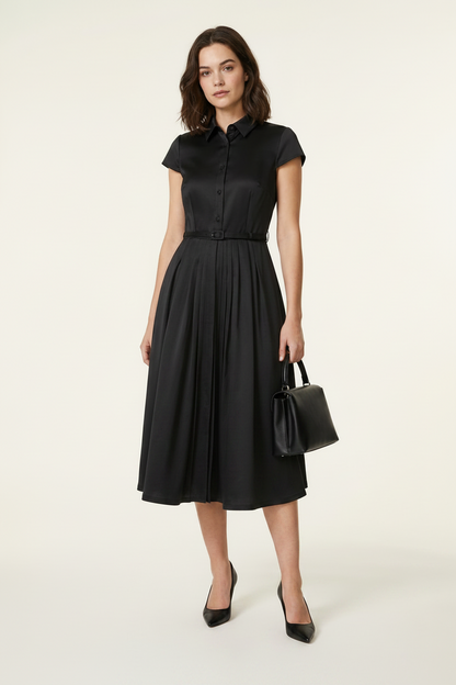 A woman modeling a black short-sleeve midi dress with a button-front bodice and flared skirt, holding a small black handbag against a plain light beige studio background.