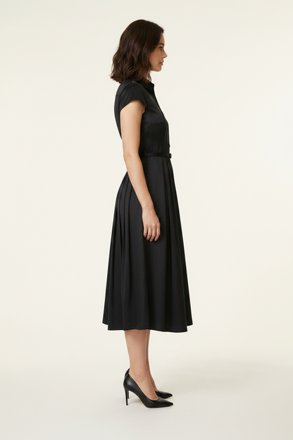 A woman modeling a black short-sleeve midi dress with a button-front bodice and flared skirt, holding a small black handbag against a plain light beige studio background.
