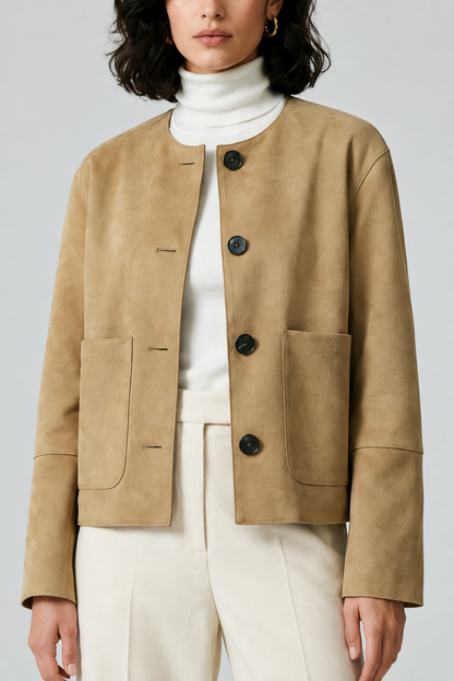 A model wearing a cropped tan suede jacket with dark buttons over a white turtleneck and cream trousers, shot against a light gray studio background.