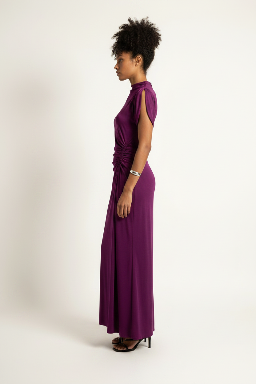 A woman modeling a sleeveless, high-neck, floor-length purple dress with a gathered waist detail, shown in profile against a plain off-white studio background.
