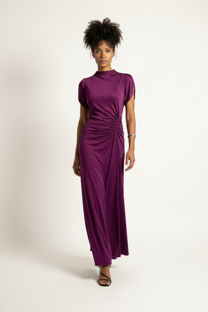 A woman modeling a high-neck, short-sleeved maxi dress in a rich purple shade, standing against a plain off-white studio background.