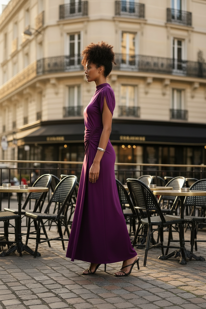 A woman stands on a cobblestone street in front of a Parisian-style building, wearing a sleeveless, high-neck, floor-length purple dress with a gathered twist detail at the waist. Black patio chairs and tables are visible in the background.