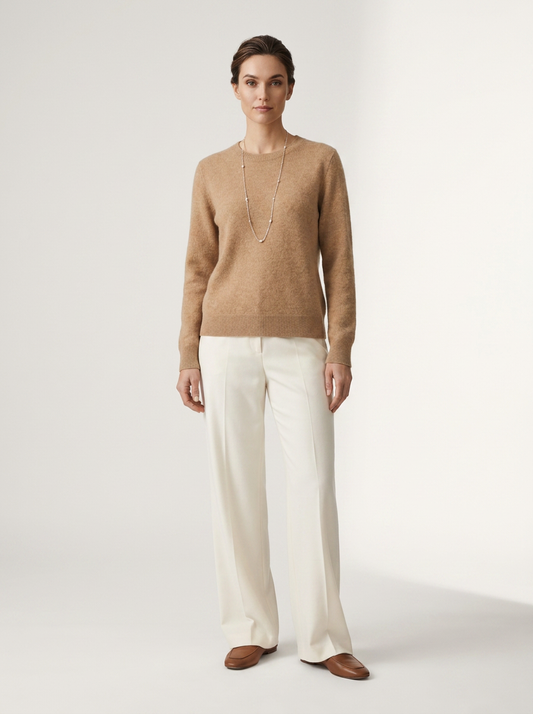 A woman stands against a plain light studio background wearing a tan knit sweater, off-white wide-leg trousers, and brown loafers, with a long silver necklace.