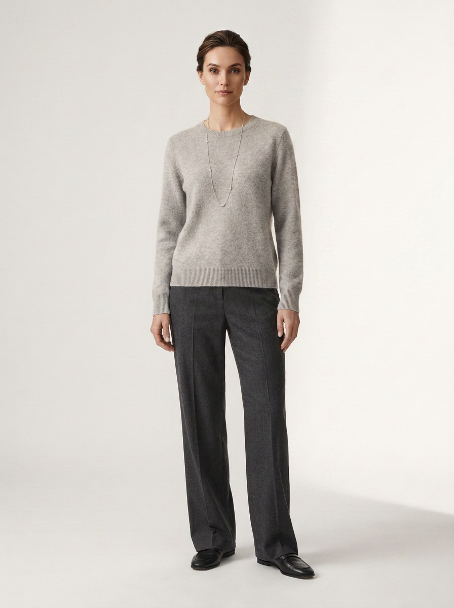 A woman stands against a plain light studio background wearing a light gray knit sweater, dark charcoal wide-leg trousers, and black shoes.