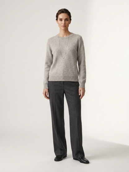 A woman stands against a plain light studio background wearing a light gray knit sweater, dark charcoal wide-leg trousers, and black shoes.