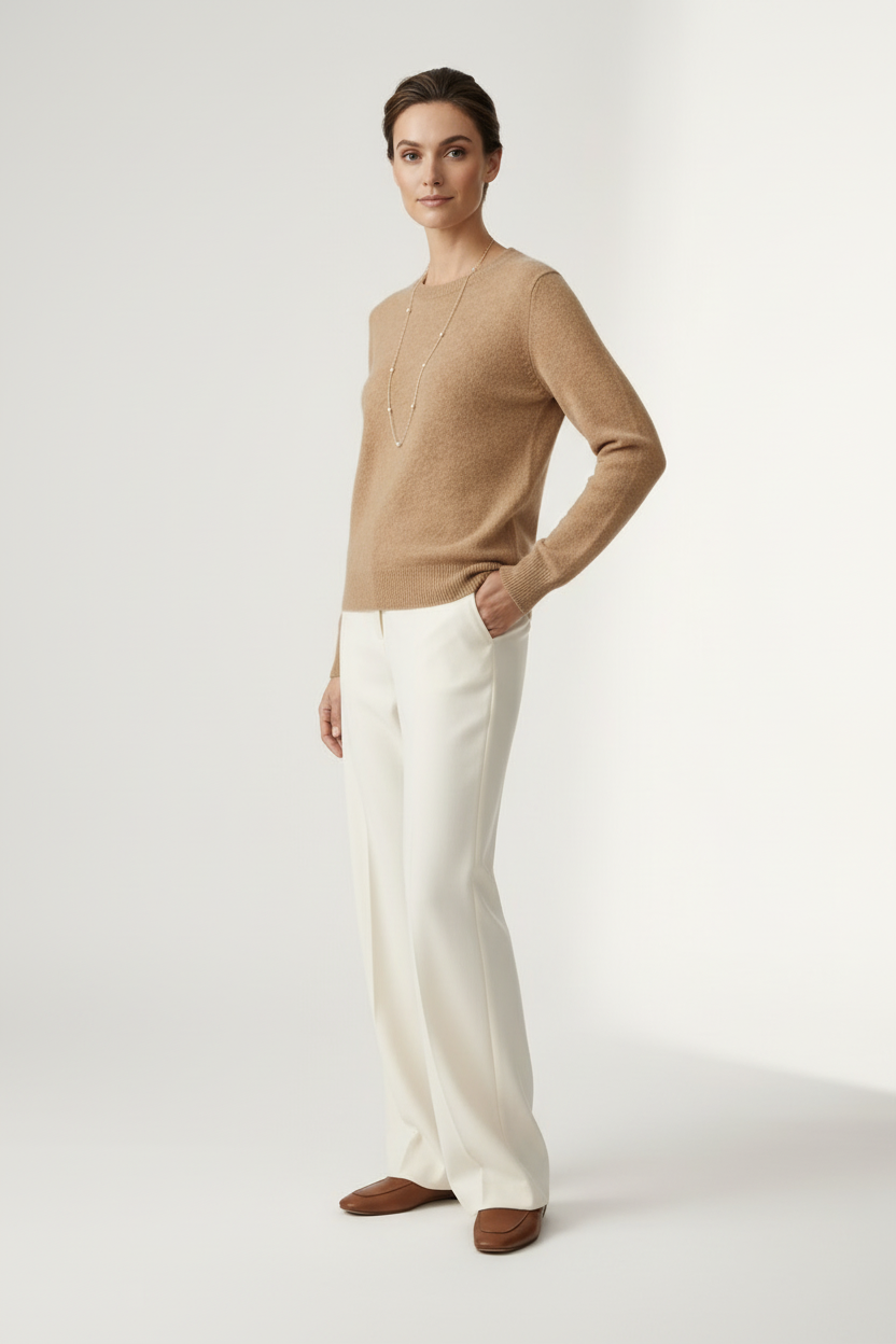 A woman stands against a plain light studio background wearing a tan knit sweater, off-white wide-leg trousers, and brown loafers, with a long silver necklace.