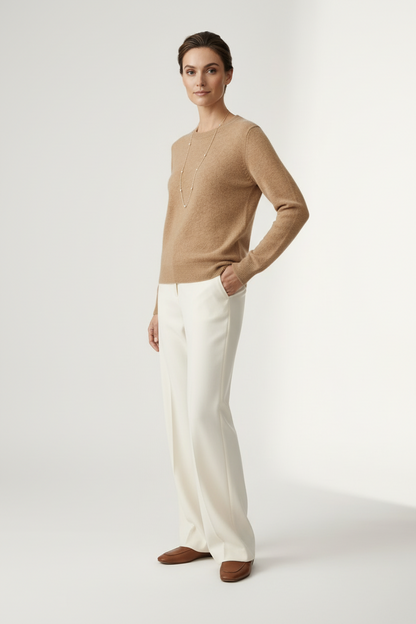 A woman stands against a plain light studio background wearing a tan knit sweater, off-white wide-leg trousers, and brown loafers, with a long silver necklace.