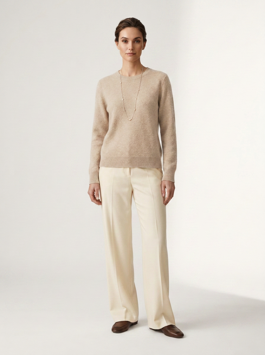 A woman stands against a plain light studio background wearing a beige knit sweater, cream wide-leg trousers, and brown loafers, with a long delicate necklace.