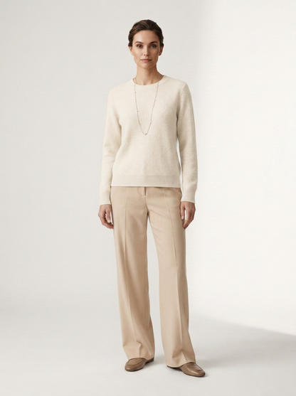 A woman stands against a plain light studio background wearing a cream knit sweater, beige wide-leg trousers, and brown loafers.