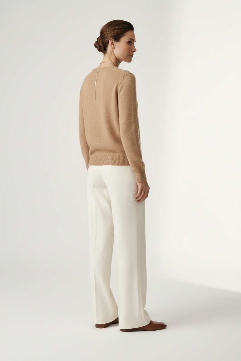 A woman stands against a plain light studio background wearing a tan knit sweater, off-white wide-leg trousers, and brown loafers, with a long silver necklace.