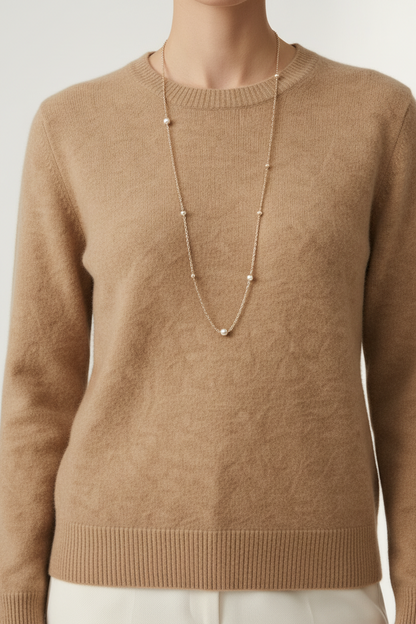 A woman stands against a plain light studio background wearing a tan knit sweater, off-white wide-leg trousers, and brown loafers, with a long silver necklace.