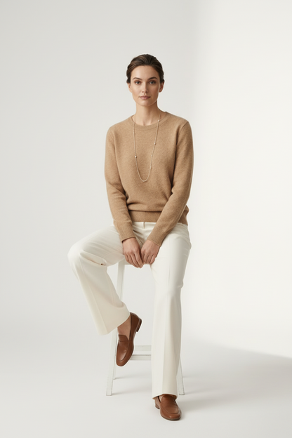 A woman stands against a plain light studio background wearing a tan knit sweater, off-white wide-leg trousers, and brown loafers, with a long silver necklace.