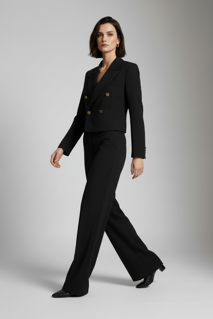 A model stands against a light gray studio background wearing a tailored black double-breasted blazer with gold buttons and matching wide-leg trousers.