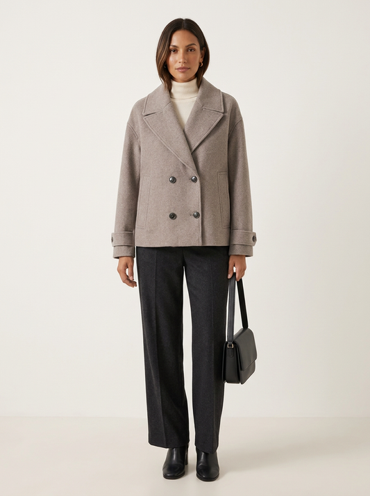 A woman stands against a plain light beige studio background wearing a double-breasted taupe wool-blend coat over a white turtleneck, dark charcoal wide-leg trousers, black ankle boots, and holding a structured black handbag.