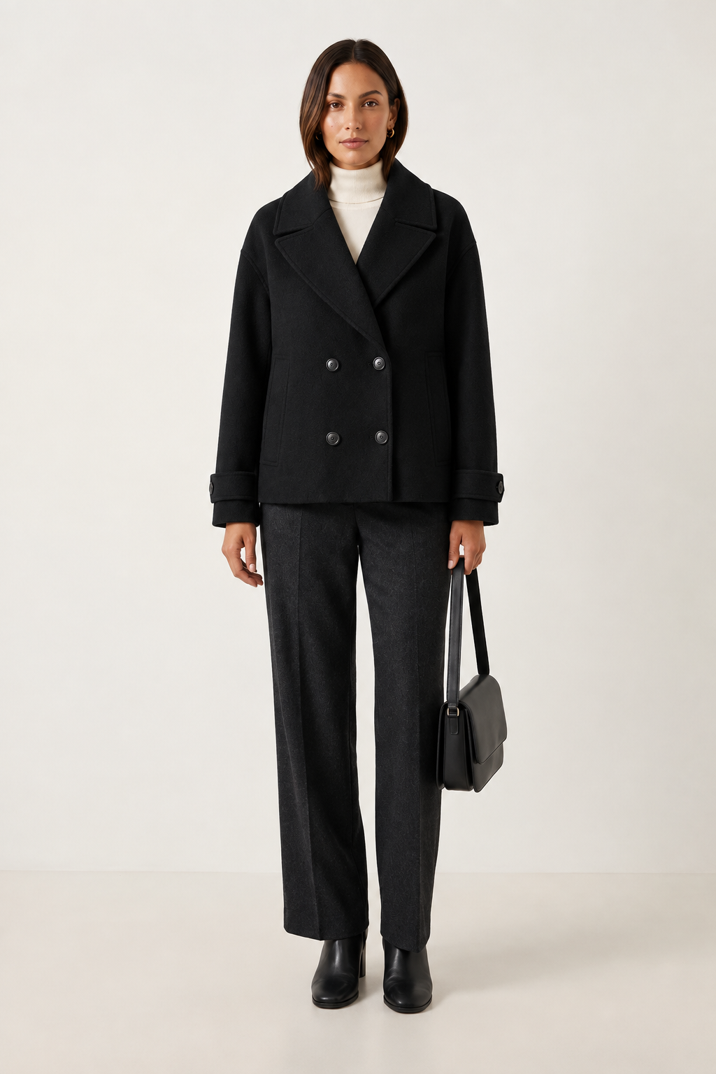 A woman stands against a plain light beige studio background wearing a double-breasted taupe wool-blend coat over a white turtleneck, dark charcoal wide-leg trousers, black ankle boots, and holding a structured black handbag.