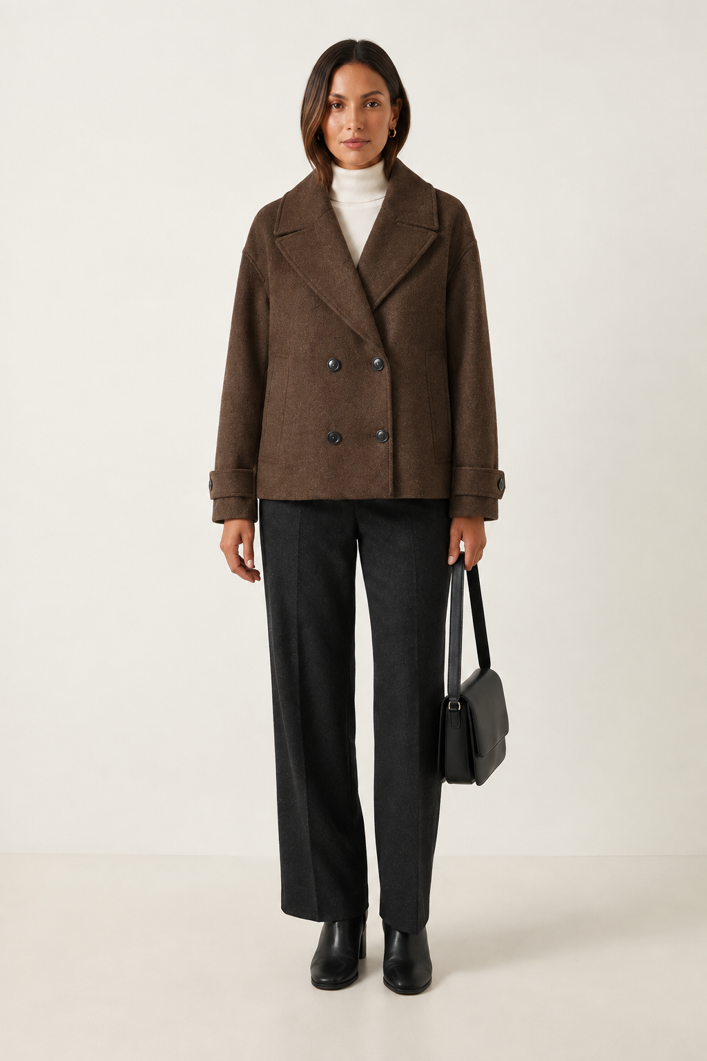 A woman stands against a plain light beige studio background wearing a double-breasted taupe wool-blend coat over a white turtleneck, dark charcoal wide-leg trousers, black ankle boots, and holding a structured black handbag.