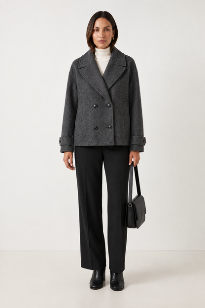 A woman stands against a plain light beige studio background wearing a double-breasted taupe wool-blend coat over a white turtleneck, dark charcoal wide-leg trousers, black ankle boots, and holding a structured black handbag.