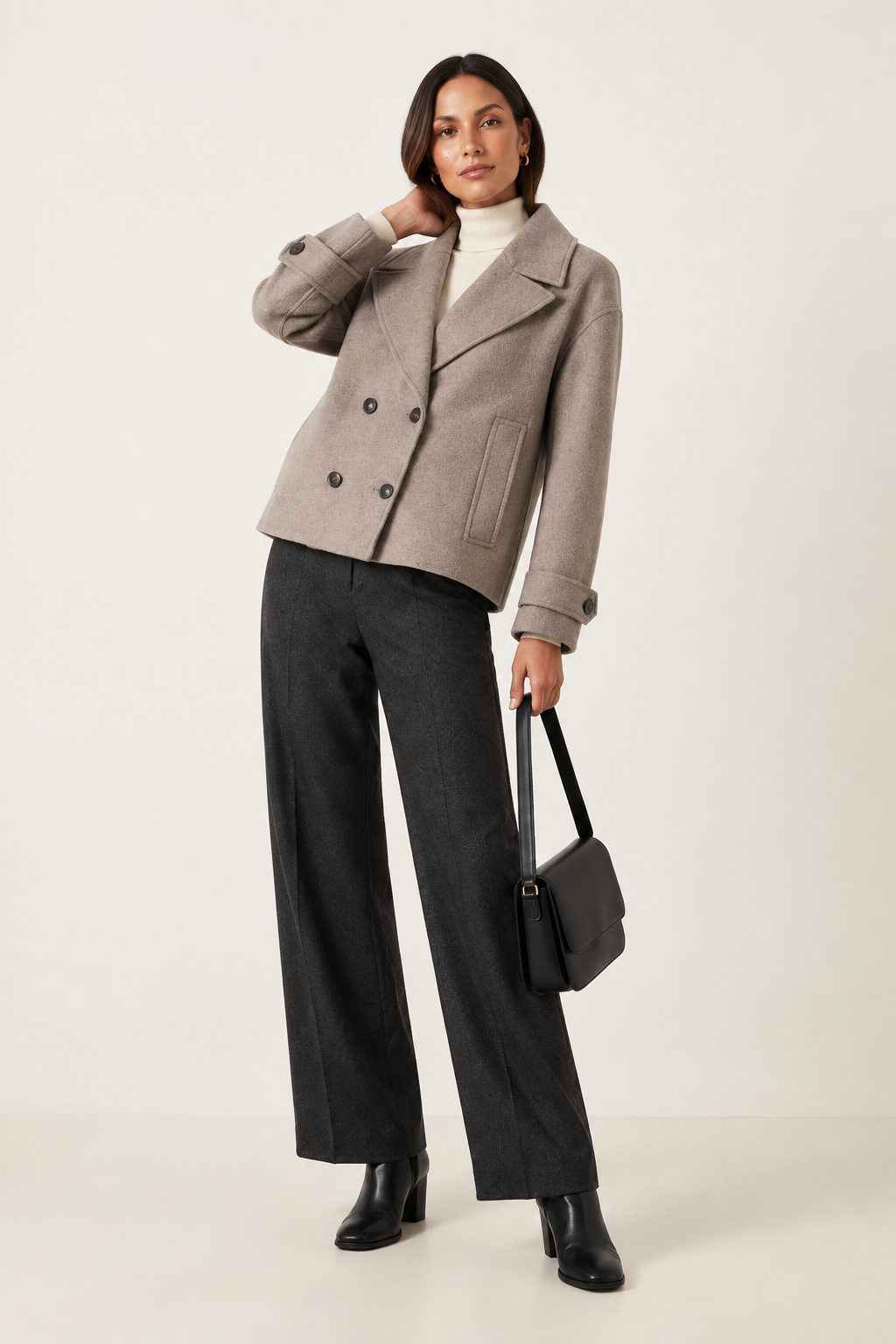 A model stands against a light beige studio background wearing a taupe double-breasted cropped jacket, dark charcoal wide-leg trousers, a white turtleneck, black ankle boots, and holding a black leather handbag.