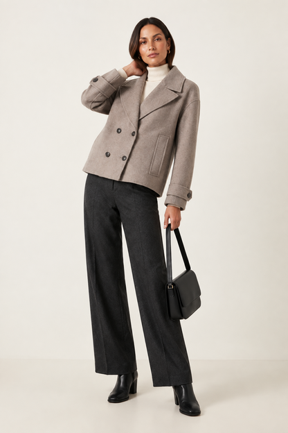 A model stands against a light beige studio background wearing a taupe double-breasted cropped jacket, dark charcoal wide-leg trousers, a white turtleneck, black ankle boots, and holding a black leather handbag.