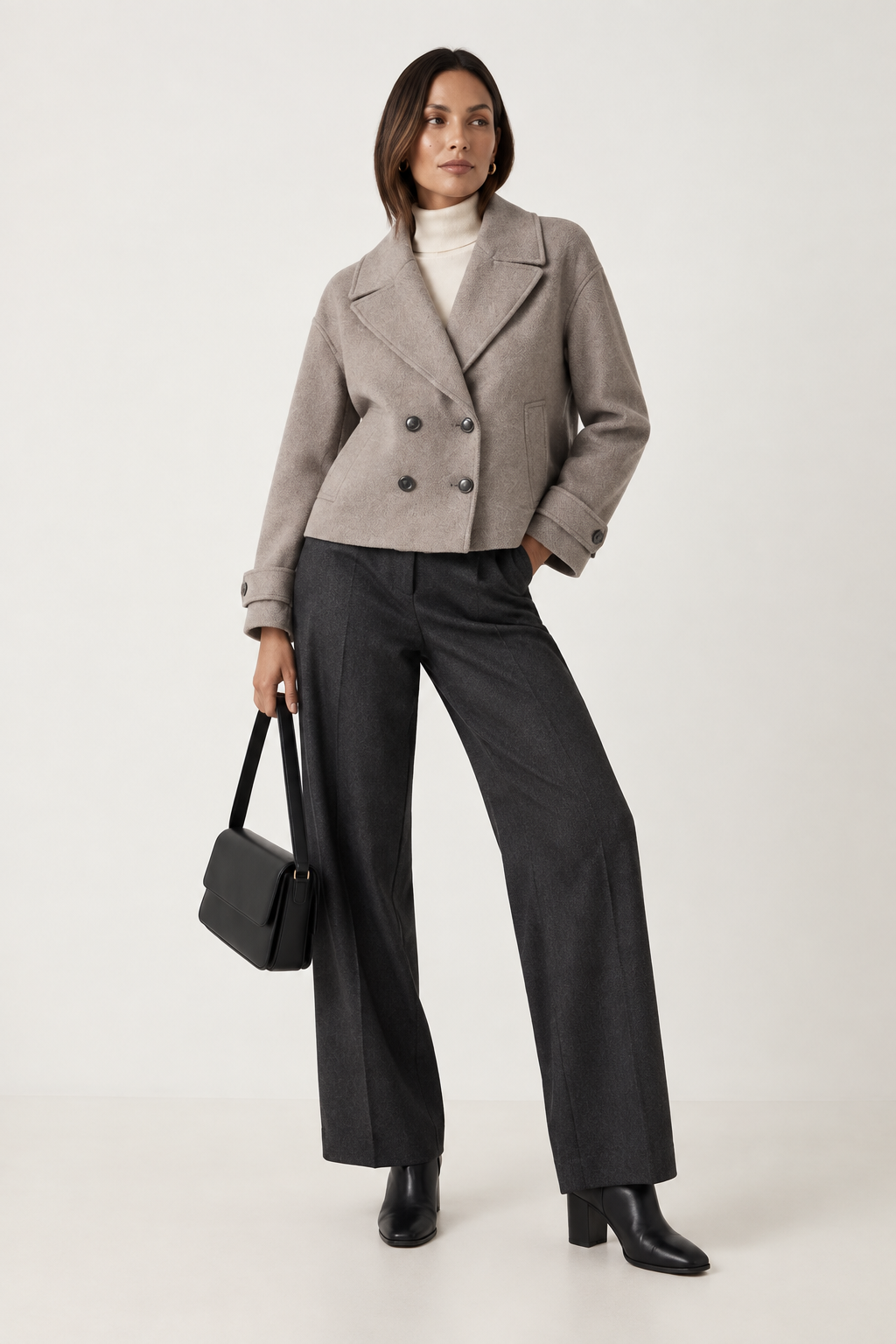 A model stands against a plain light background wearing a light taupe double-breasted cropped coat over a white turtleneck, dark charcoal wide-leg trousers, black ankle boots, and holding a black handbag.