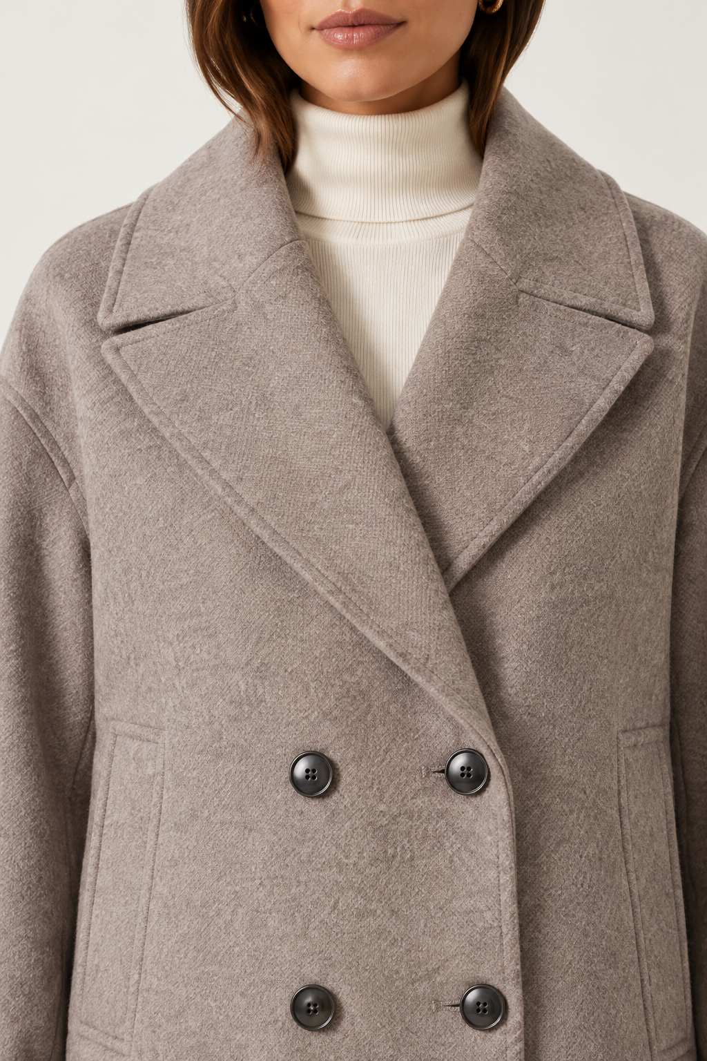 A close-up of a model wearing a double-breasted taupe wool-blend coat over a cream turtleneck sweater.