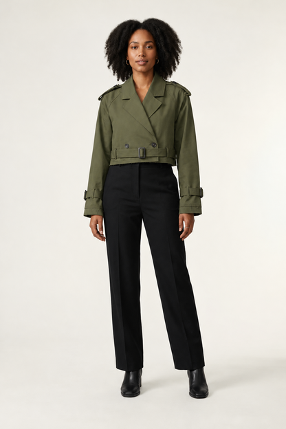 A model stands against a plain light background wearing a beige trench-style jacket with shoulder epaulets and a belt, paired with black trousers and black ankle boots.