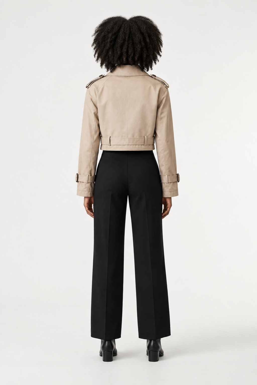A model stands against a plain light background wearing a beige trench-style jacket with shoulder epaulets and a belt, paired with black trousers and black ankle boots.