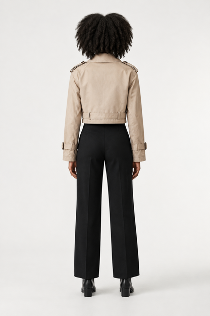 A model stands against a plain light background wearing a beige trench-style jacket with shoulder epaulets and a belt, paired with black trousers and black ankle boots.