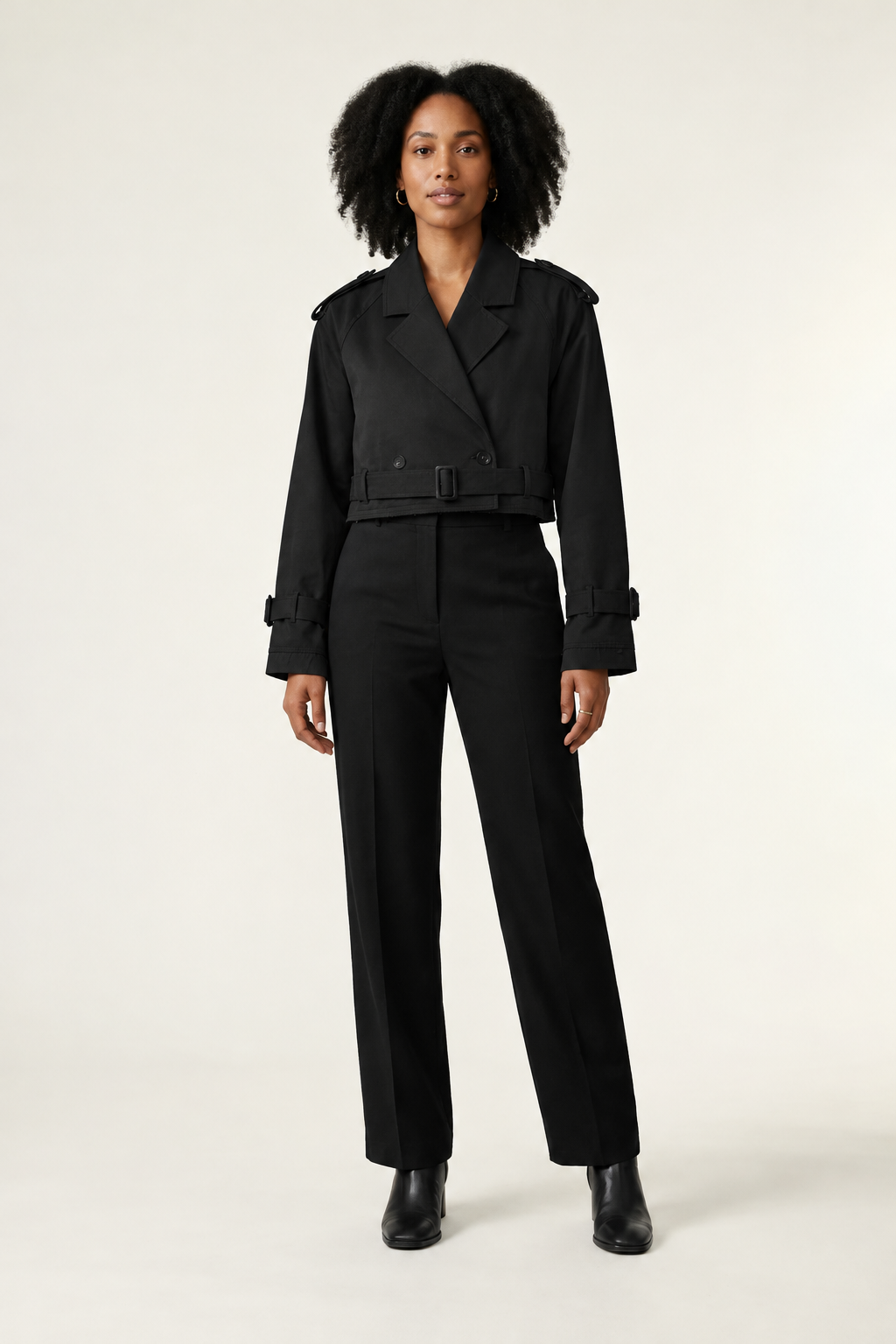 A model stands against a plain light background wearing a beige trench-style jacket with shoulder epaulets and a belt, paired with black trousers and black ankle boots.