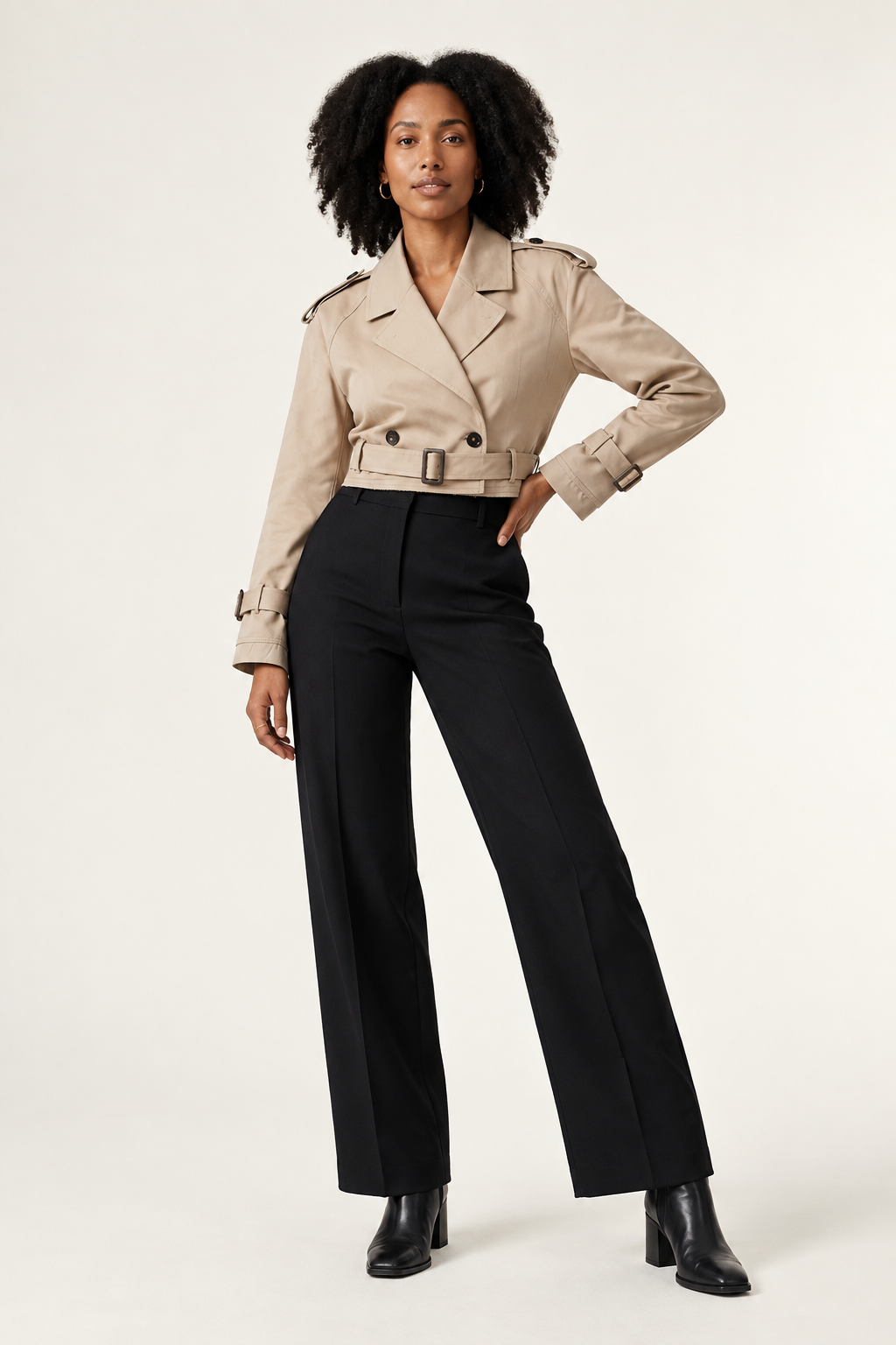 A model stands against a plain light background wearing a beige trench-style jacket with shoulder epaulets and a belt, paired with black trousers and black ankle boots.