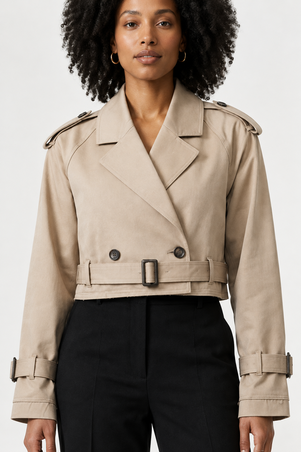 A model stands against a plain light background wearing a beige trench-style jacket with shoulder epaulets and a belt, paired with black trousers and black ankle boots.