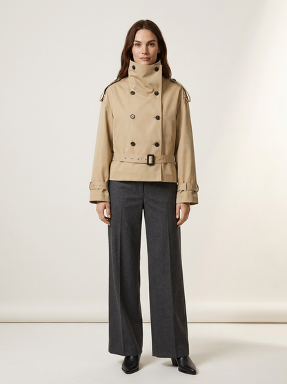 A model stands against a plain off-white studio background wearing a beige double-breasted trench coat with a high collar and dark charcoal wide-leg trousers.