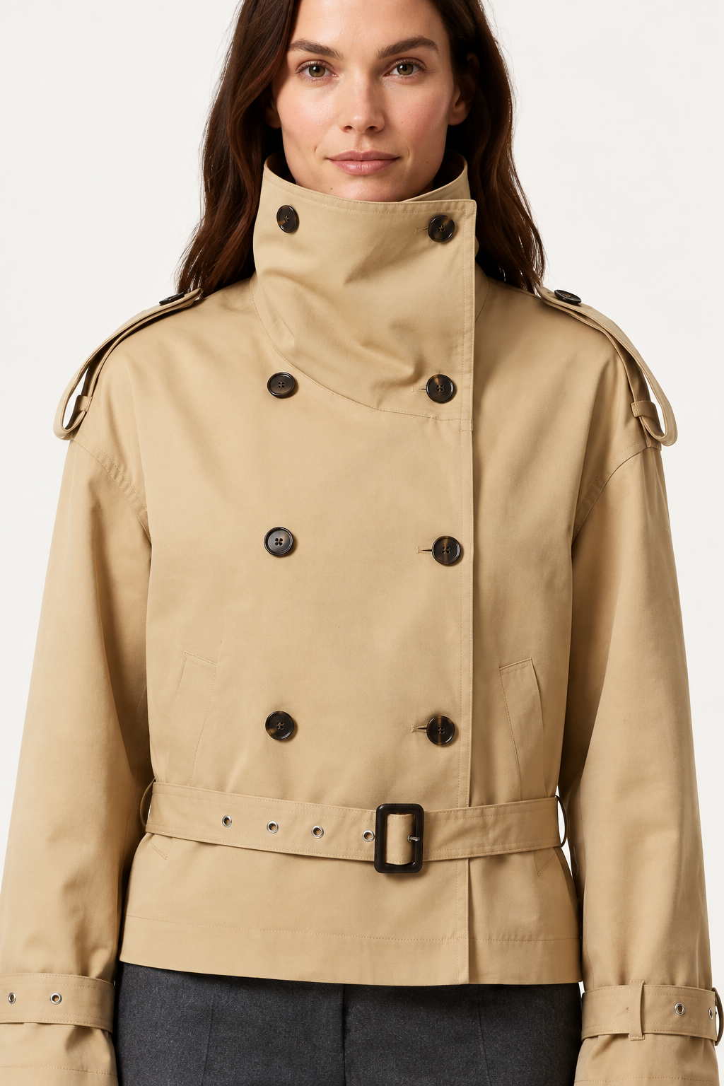 A woman wearing a beige double-breasted trench coat with a high collar and matching belt, photographed against a plain light background.