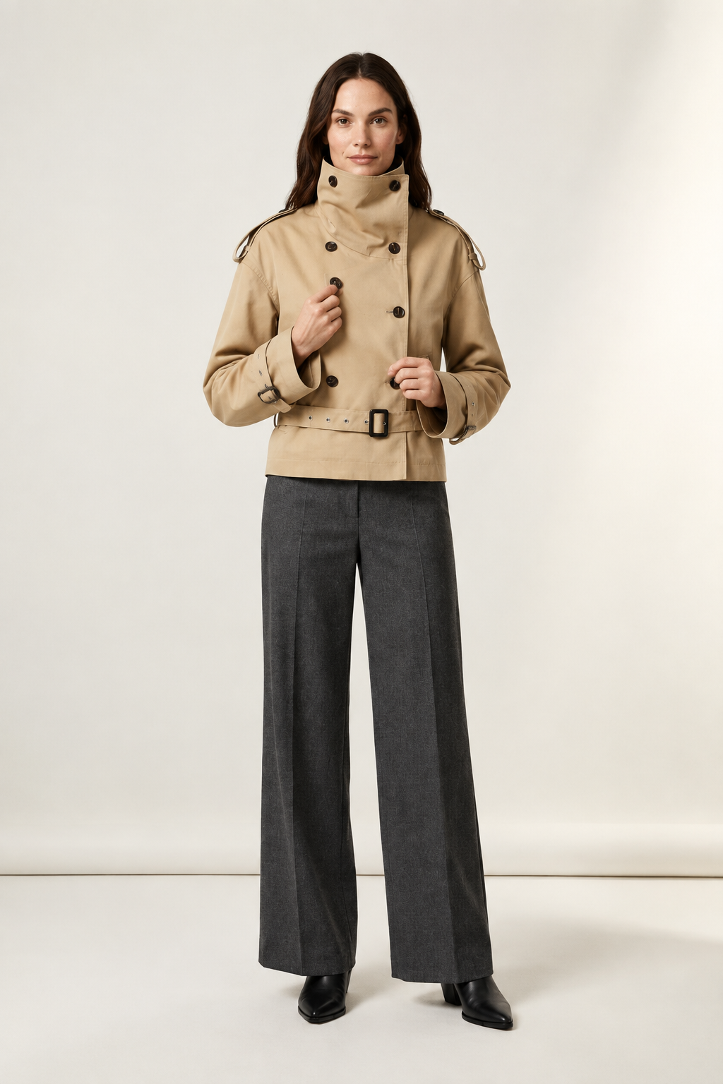 A model stands against a plain light beige studio background wearing a beige double-breasted trench jacket with a high collar and dark buttons, paired with dark gray wide-leg trousers and black pointed shoes.