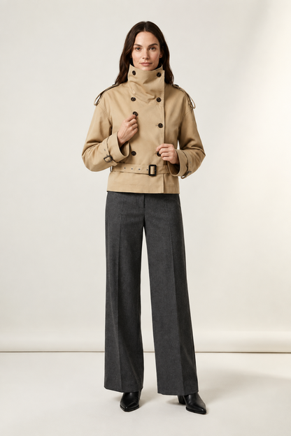 A model stands against a plain light beige studio background wearing a beige double-breasted trench jacket with a high collar and dark buttons, paired with dark gray wide-leg trousers and black pointed shoes.