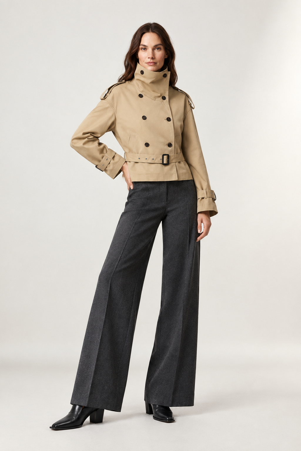 A model stands against a light gray studio background wearing a beige double-breasted trench jacket with a belted waist, dark charcoal wide-leg trousers, and black pointed-toe heels.