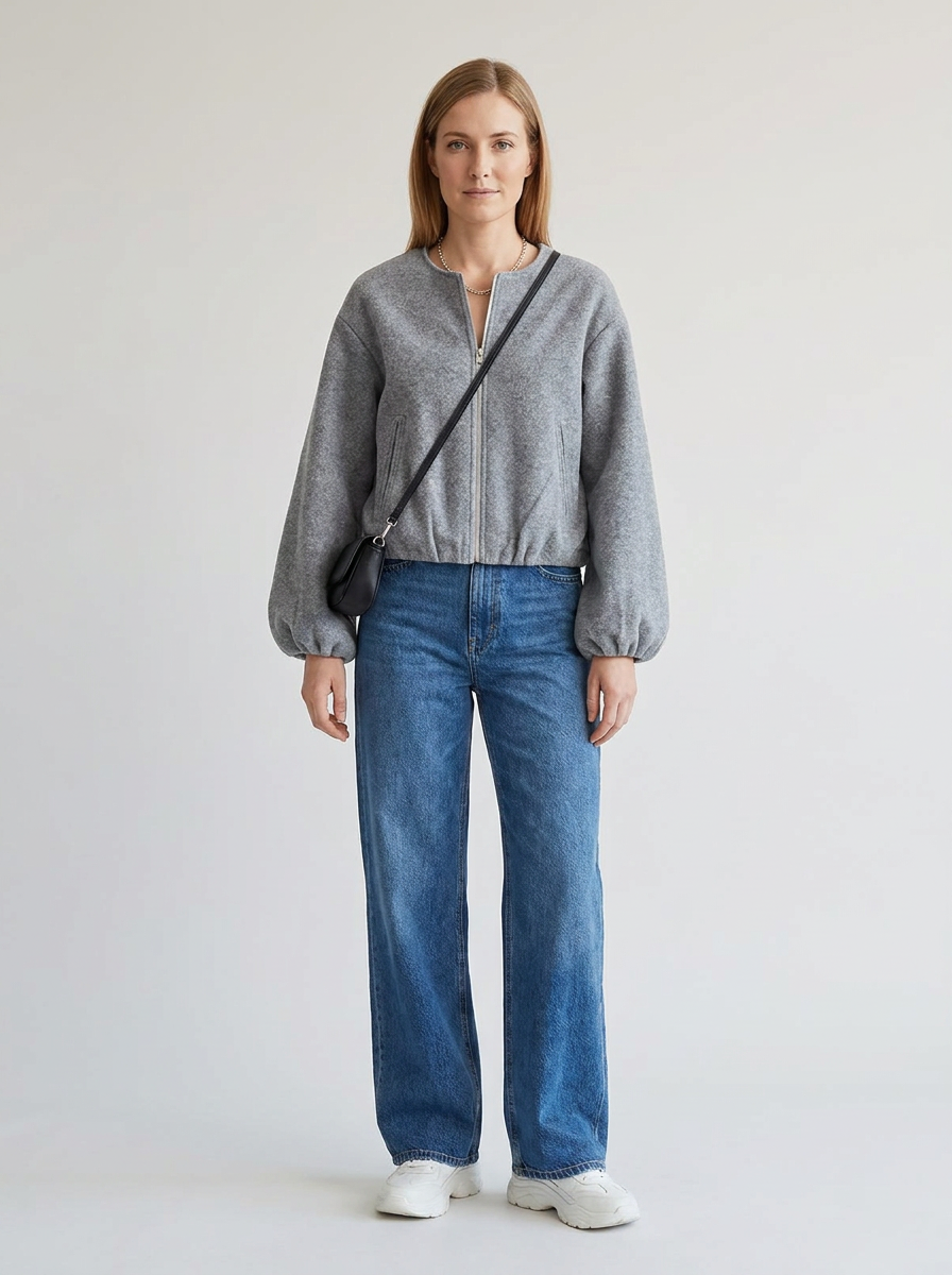 A woman stands against a plain light gray studio background wearing a gray cropped zip-front sweater, blue wide-leg jeans, white sneakers, and a black crossbody bag.