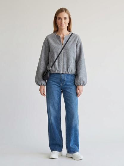 A woman stands against a plain light gray studio background wearing a gray cropped zip-front sweater, blue wide-leg jeans, white sneakers, and a black crossbody bag.