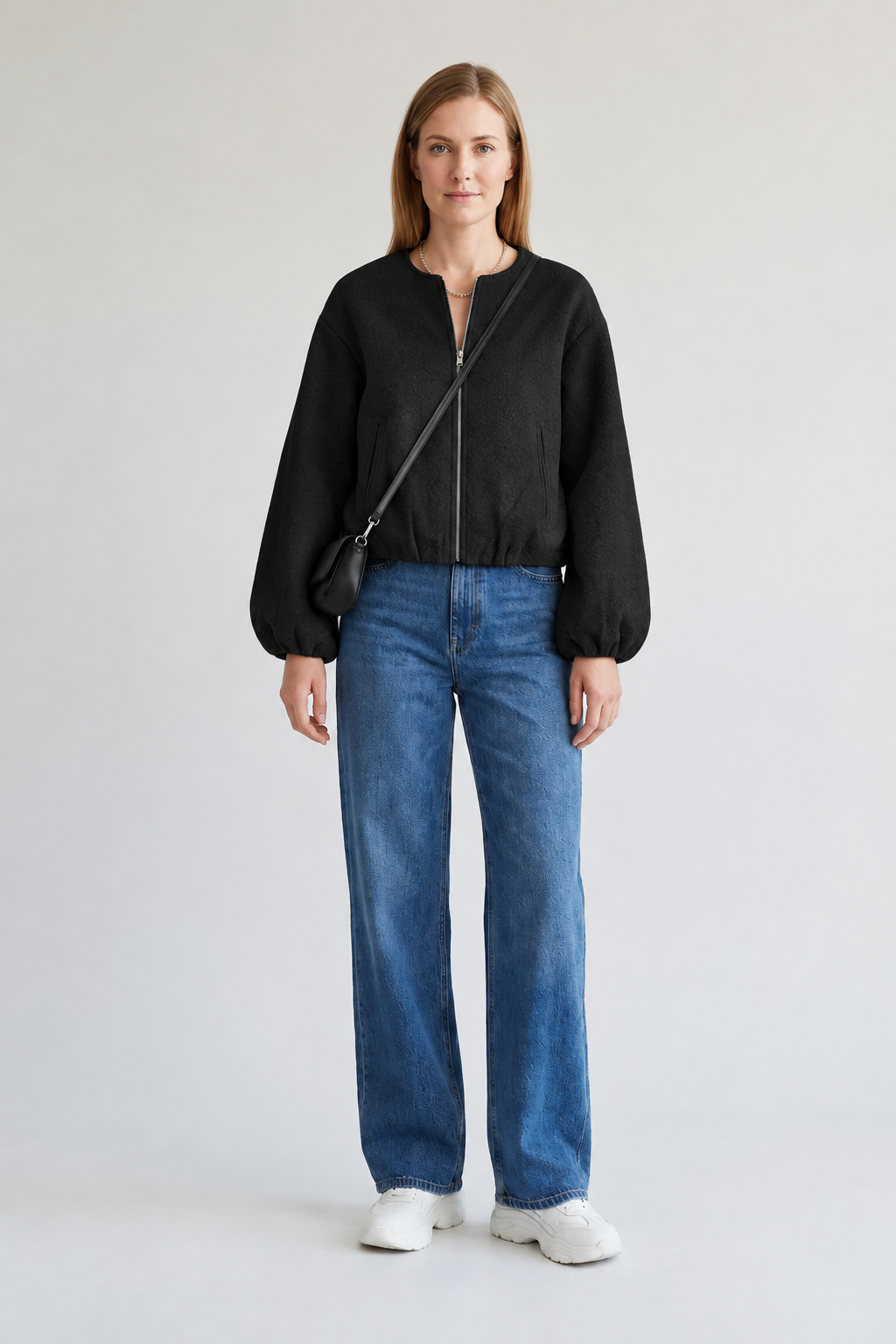 A woman stands against a plain light gray studio background wearing a gray cropped zip-front sweater, blue wide-leg jeans, white sneakers, and a black crossbody bag.