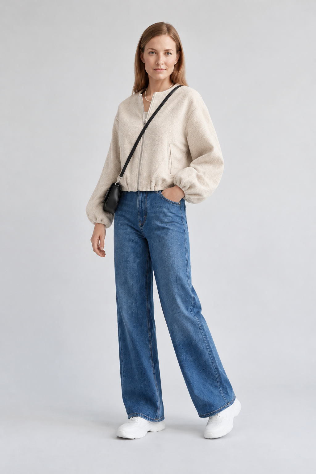 A woman stands against a light gray studio background wearing a cream sherpa zip-up jacket, blue wide-leg jeans, white sneakers, and a small black crossbody bag.