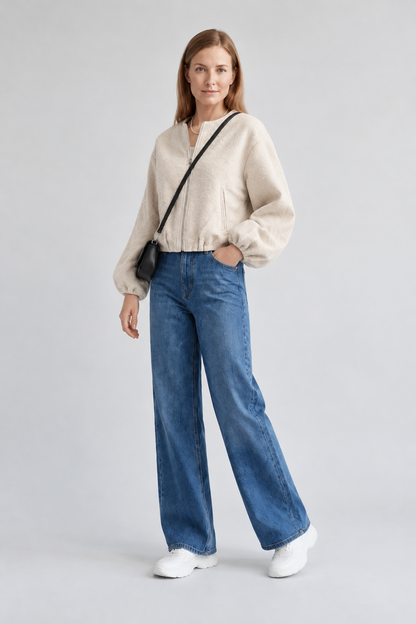 A woman stands against a light gray studio background wearing a cream sherpa zip-up jacket, blue wide-leg jeans, white sneakers, and a small black crossbody bag.