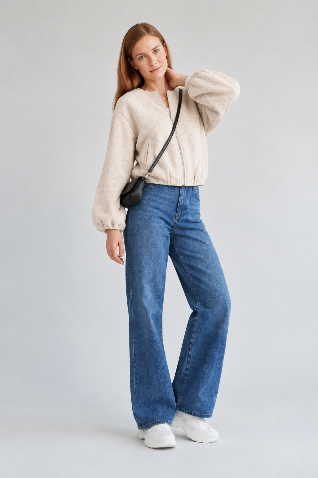 A woman stands against a light gray studio background wearing a cream sherpa zip-up jacket, blue wide-leg jeans, white sneakers, and a small black crossbody bag.