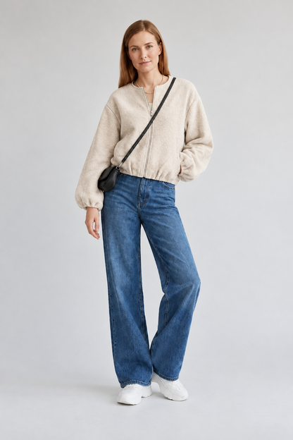 A woman stands against a light gray studio background wearing a cream sherpa zip-up jacket, blue wide-leg jeans, white sneakers, and a small black crossbody bag.