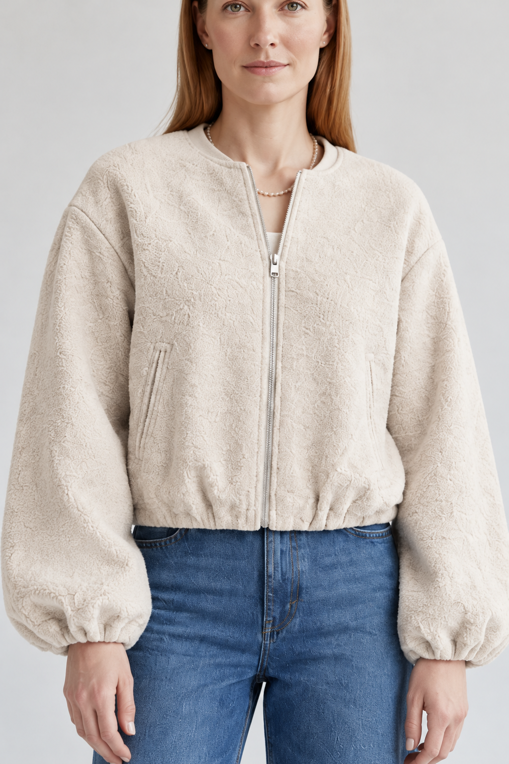 A woman stands against a light gray studio background wearing a cream sherpa zip-up jacket, blue wide-leg jeans, white sneakers, and a small black crossbody bag.